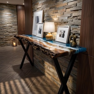 Può includere: Un tavolo console con un piano in legno a bordo vivo e un inserto in resina blu e bianca, sostenuto da gambe in metallo nero a forma di X. La superficie ospita una lampada da tavolo, tre stampe incorniciate e una piccola scultura in bronzo.