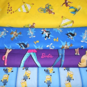 May include: A stack of colorful fabric rolls featuring cartoon characters and designs. The top roll is yellow with a circus theme. The second roll is blue with Looney Tunes characters. The third roll is purple with a Barbie design. The bottom roll is light blue with Minions.