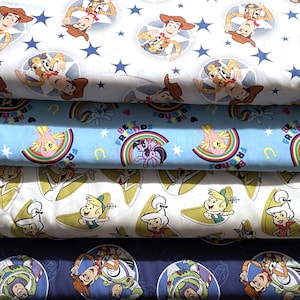 May include: A stack of colorful fabric featuring cartoon characters. The top layer has a white background with Woody from Toy Story, surrounded by blue stars. Below is a light blue fabric with My Little Pony characters and the text "BEST FRIENDS".