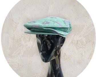 May include: A light green flat cap featuring a repeating pattern of purple abstract shapes. The cap has a structured brim and a rounded crown, displayed on a black mannequin head. The fabric appears to have a soft texture with a consistent color.