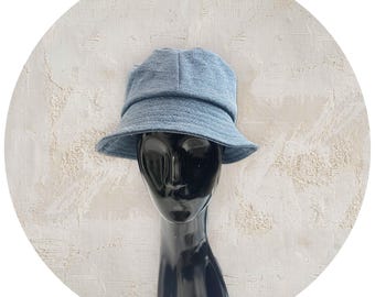 May include: A denim blue bucket hat with a rounded crown and a downward-sloping brim. The hat is displayed on a black mannequin head against a textured white background. The hat's fabric appears soft and casual, suitable for everyday wear.