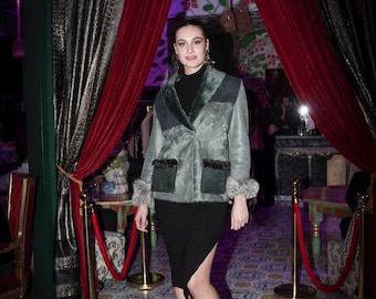 May include: A sage green jacket with a wide collar, dark green suede-like panels, and two front pockets with dark fur trim. The jacket features matching fur cuffs at the sleeves and is worn over a black mock neck top and a black midi skirt with a side slit.