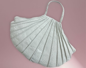 May include: A white, seashell-shaped handbag with a braided handle. The bag has a textured surface and a pleated design, with radiating lines that mimic the shape of a seashell. The background is a solid pink.