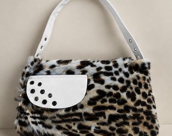 May include: A shoulder bag featuring a faux fur exterior with a brown, black, and white leopard print pattern. The bag includes a white strap and a white semi-circular front pocket flap accented with five black circular metal grommets.
