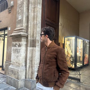 May include: A brown suede bomber jacket with a ribbed collar and cuffs. The jacket is paired with light gray shorts and sunglasses. The person is standing outside a building with a stone pillar and a wooden door.