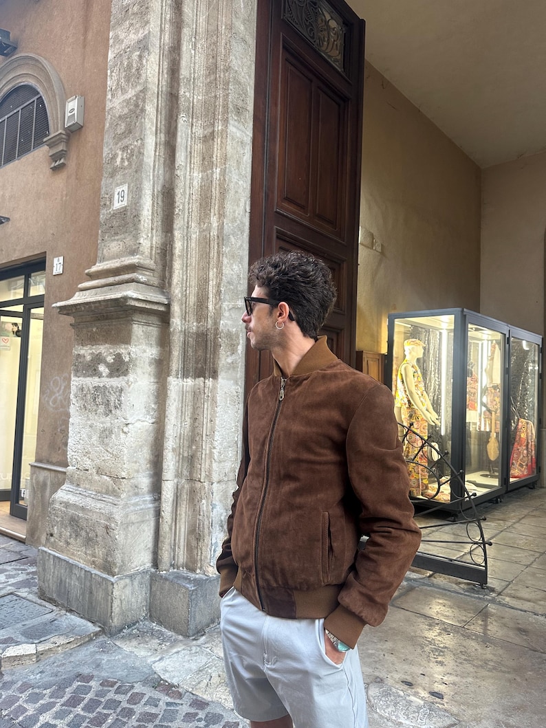 May include: A brown suede bomber jacket with a ribbed collar and cuffs. The jacket is paired with light gray shorts and sunglasses. The person is standing outside a building with a stone pillar and a wooden door.