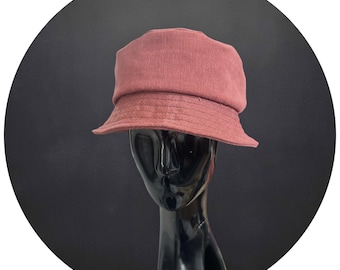 May include: A dusty rose bucket hat with a structured crown and a downward-sloping brim. The hat is made of a textured fabric and is displayed on a black mannequin head against a dark background. This hat is a stylish accessory.