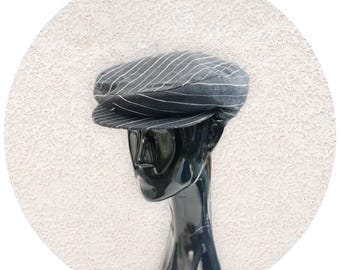May include: A gray newsboy cap with white pinstripes. The cap is displayed on a black mannequin head against a textured white background. The cap has a rounded crown and a short, stiff brim.