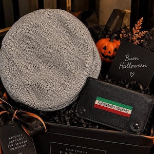 Puede incluir: Una gorra plana de tweed gris y una cartera de cuero negro con un parche rectangular con los colores de la bandera italiana y el texto FERDINANDO PATERMO, presentados en una caja de regalo con una calabaza y una tarjeta Buon Halloween.