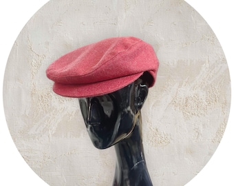 May include: A red newsboy cap with a rounded crown and a small, stiff brim. The cap is made of a textured fabric and is displayed on a black mannequin head against a white textured background.
