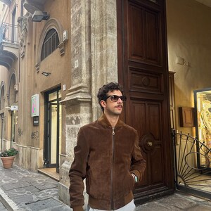 May include: A brown suede bomber jacket with a zipper closure. The jacket has a ribbed collar and cuffs. The person is wearing sunglasses and gray pants. The background includes a building with a wooden door.