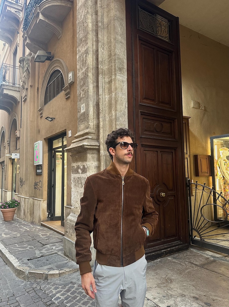 May include: A brown suede bomber jacket with a zipper closure. The jacket has a ribbed collar and cuffs. The person is wearing sunglasses and gray pants. The background includes a building with a wooden door.