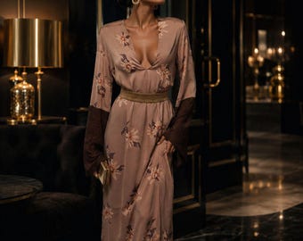 May include: A long-sleeved, floor-length dress in a muted mauve tone featuring a floral pattern in shades of cream and brown. The garment includes a deep V-neckline, a wide gold-toned waistband, and dark brown contrasting cuffs on the sleeves.