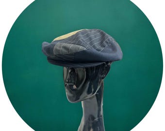 May include: A blue and gray patterned newsboy cap with a slightly curved brim. The cap is on a black mannequin head against a green background.