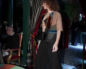 May include: A cropped jacket featuring a patchwork design of tan, brown, and black panels with a metallic gold front closure, paired with high-waisted black wide-leg trousers. The jacket has short sleeves and a collarless, open-front construction.