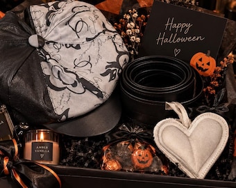 May include: A gift box containing a black and grey newsboy cap with script patterns, a coiled black leather belt, a candle labeled Amber Vanilla, a heart-shaped grey fabric ornament, and a card with the text Happy Halloween and a pumpkin icon.