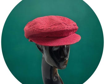 May include: A red perforated newsboy cap with a rounded crown and a short, stiff brim. The cap is displayed on a black mannequin head against a teal green background. The hat's texture appears soft and breathable.