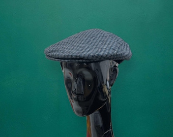 May include: A grey and black houndstooth patterned flat cap featuring a structured crown and a short, stiff brim. The cap is displayed on a glossy black mannequin head, showcasing the textured fabric weave and the classic low-profile silhouette.