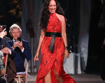 May include: A sleeveless red floor-length dress featuring a black abstract pattern, a high halter neckline, and a wide black leather-like waist belt. The dress has a front slit that reveals the legs and is paired with metallic gold strappy sandals.