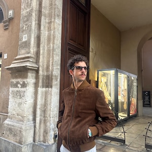 May include: A brown suede bomber jacket with a zip closure and ribbed cuffs and hem. The jacket has a brown collar and a light brown lining. The person is wearing sunglasses and light gray pants.