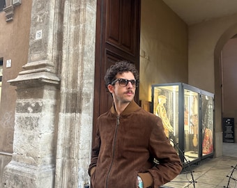 May include: A brown suede bomber jacket with a zip closure and ribbed cuffs and hem. The jacket has a brown collar and a light brown lining. The person is wearing sunglasses and light gray pants.