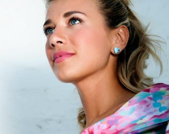 May include: A bright turquoise heart-shaped stud earring worn on the earlobe. The earring features a small, centered dark red accent detail. The piece is crafted with a smooth finish and is positioned to be clearly visible against the skin.