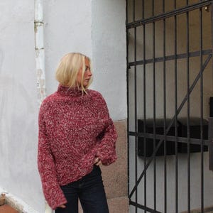 May include: A woman wearing a red and white fuzzy sweater with a high neck and long sleeves. She is wearing dark blue jeans and is standing in front of a metal gate.