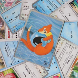 May include: A close-up of a Pokemon card featuring a cartoon orange fox holding a blue and black sphere. The card is surrounded by other Pokemon cards, including cards with the text "Surf", "Aqua Slash", "Permeating", "Icicle Missile", "Flap", "Nosedive", "Vise Grip", "Call for Family", "Egg Rolling", "Pound", and "Mise en Place".