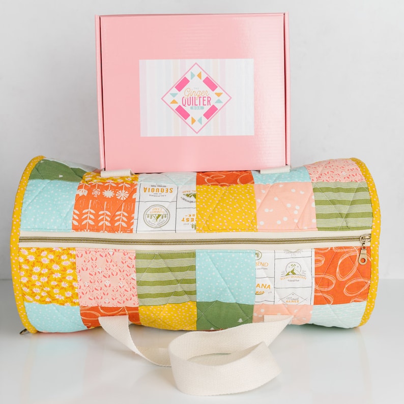 May include: A pink box with the text "Ginger Quilter" on it, sitting on top of a quilted fabric duffel bag with a white strap. The bag is made of patchwork fabric with a variety of colors and patterns, including orange, yellow, green, blue, and pink.