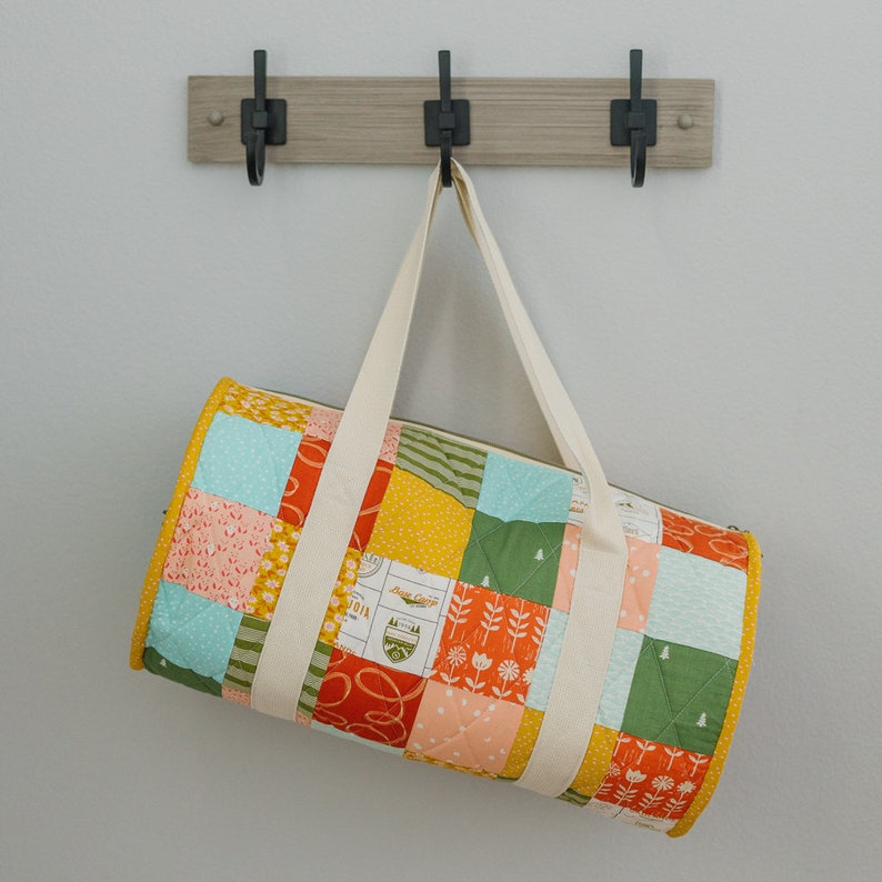 May include: A patchwork style duffel bag with a cream canvas strap. The bag is made of a variety of fabrics in shades of green, yellow, orange, pink, and blue. The bag has a quilted design and features a variety of patterns, including floral, geometric, and plaid.