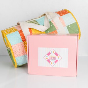 May include: A pink box with a white label that says "Ginger Quilter" on it. The box is next to a patchwork fabric tote bag with a white strap.