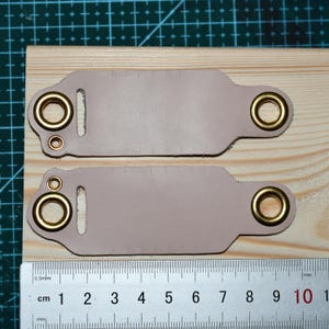 May include: Two light mauve leather pieces with brass eyelets, placed on a wooden surface. A ruler with centimeter and millimeter markings is visible, indicating measurements. The leather pieces have a rectangular shape with rounded edges.