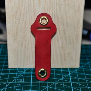 May include: A red leather key fob with gold-colored grommets. The key fob has a rectangular shape with a rounded top and bottom. The key fob is shown against a light-colored wooden surface.