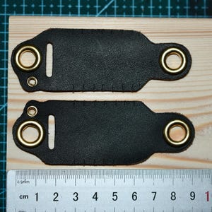 May include: Two black leather pieces with brass eyelets and stitching, laid on a wooden surface. Each piece has a rectangular shape with rounded ends and a slot. A ruler with centimeter markings is visible.