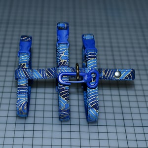 May include: A blue and gold patterned pet harness with adjustable straps and plastic buckles. The harness features a metal ring and a blue plastic clasp for leash attachment. The design includes a repeating wave-like pattern in gold against a blue background.