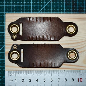 May include: Two brown leather straps with brass eyelets and stitched edges. Each strap has a rectangular shape with a rounded end and a slot for attachment. The straps are placed on a wooden surface with a ruler.