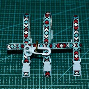 May include: A turquoise dog harness with a patterned design in red, white, and black. The harness features plastic buckles and a metal ring for leash attachment. The design is symmetrical and the harness is laid out on a green grid.
