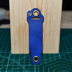 May include: A cobalt blue leather strap with gold-colored grommets. The strap is rectangular with a rounded top and a small hole. The background includes a wooden block and a green cutting mat.