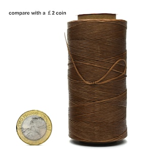 Può includere: Un rocchetto di filo cerato marrone per la lavorazione del cuoio, confrontato con una moneta da 2 sterline per riferimento alle dimensioni. Il testo "compare with a £2 coin" è visibile nell'immagine.