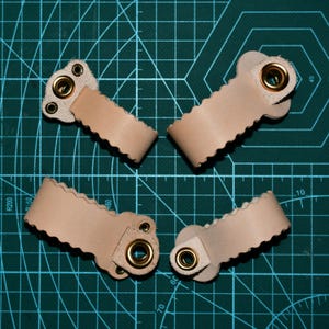 May include: Four beige leather straps with scalloped edges and brass eyelets. Each strap has a rectangular base with multiple holes and a rounded end with a brass eyelet. The straps are arranged on a green cutting mat.