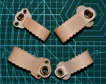 May include: Four beige leather straps with scalloped edges and brass eyelets. Each strap has a rectangular base with multiple holes and a rounded end with a brass eyelet. The straps are arranged on a green cutting mat.