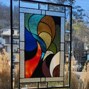 May include: Stained glass window panel with a black metal frame. The design features abstract shapes in shades of blue, orange, brown, green, and yellow. The panel is hanging outside, with a chain and the sky visible in the background.