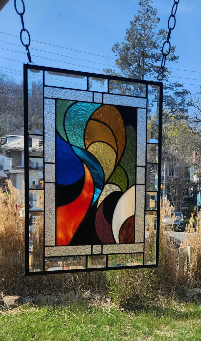 May include: Stained glass window panel with a black metal frame. The design features abstract shapes in shades of blue, orange, brown, green, and yellow. The panel is hanging outside, with a chain and the sky visible in the background.