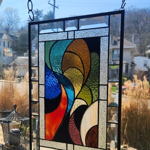 May include: A rectangular stained glass panel with a black metal frame. The design features abstract shapes in shades of blue, orange, green, brown, and white. The panel is suspended by a chain, showcasing a vibrant and colourful artwork.