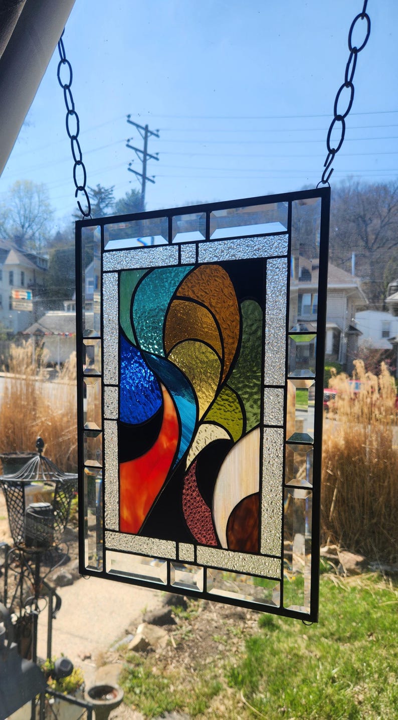 May include: A rectangular stained glass panel with a black metal frame. The design features abstract shapes in shades of blue, orange, green, brown, and white. The panel is suspended by a chain, showcasing a vibrant and colourful artwork.