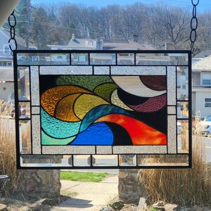 May include: A rectangular stained glass panel with a black metal frame. The design features abstract shapes in shades of blue, green, yellow, orange, and brown against a black background. The panel is suspended from a chain.