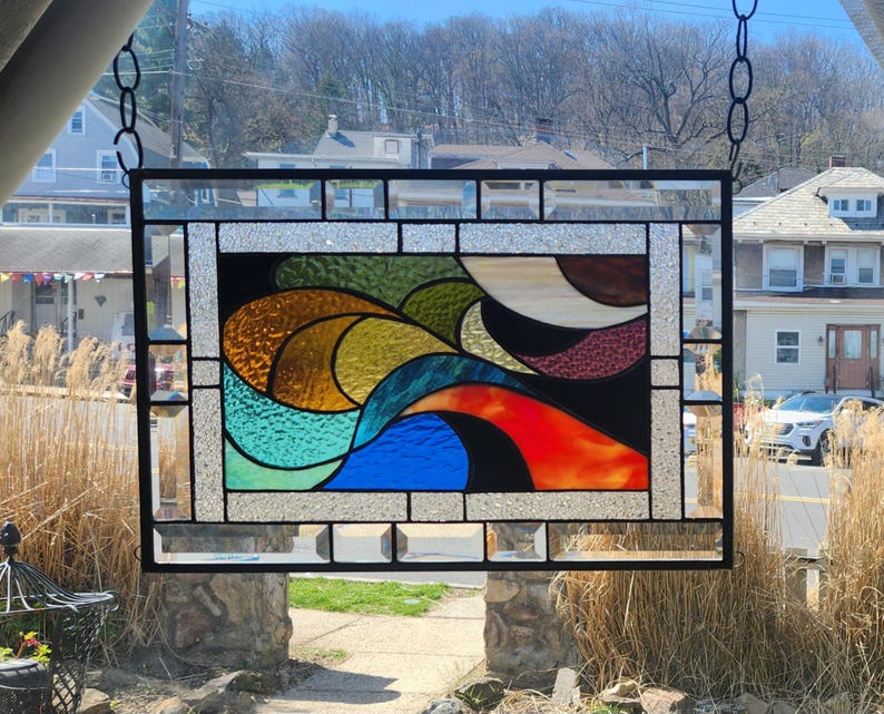 May include: A rectangular stained glass panel with a black metal frame. The design features abstract shapes in shades of blue, green, yellow, orange, and brown against a black background. The panel is suspended from a chain.