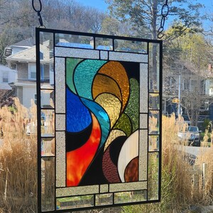 May include: A rectangular stained glass panel with a black metal frame. The design features abstract shapes in shades of blue, orange, yellow, green, and brown. The panel is hanging from a chain in front of a window, with a view of trees and houses.