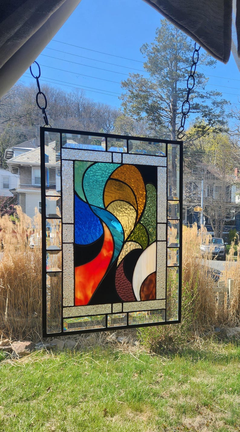 May include: A rectangular stained glass panel with a black metal frame. The design features abstract shapes in shades of blue, orange, yellow, green, and brown. The panel is hanging from a chain in front of a window, with a view of trees and houses.