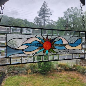 May include: Rectangular stained glass panel featuring a sun design with a red center and colorful rays. The sun is surrounded by blue and white wave-like patterns. The panel is framed in black and suspended by chains.
